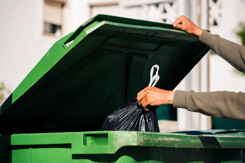 Hand throwing away black bag of rubbish in green bin