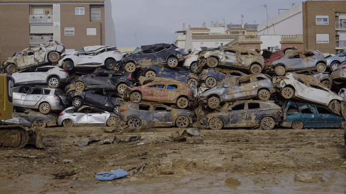 A large stack of mud-covered cars sits in a muddy lot after severe flooding in Valencia, with residential buildings visible in the background