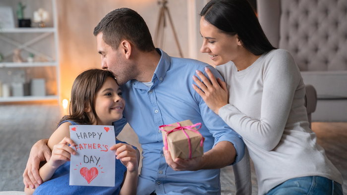 A young girl presenting a handmade Father’s Day card and gift to her smiling dad, while her mother looks on affectionately