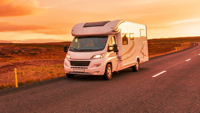 A modern white motorhome driving on an open road at sunset, surrounded by scenic countryside.