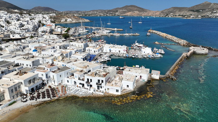 Aerial view of the picturesque harbour and whitewashed buildings of Paros island, Greece, with boats moored in clear blue water and rolling hills in the background.