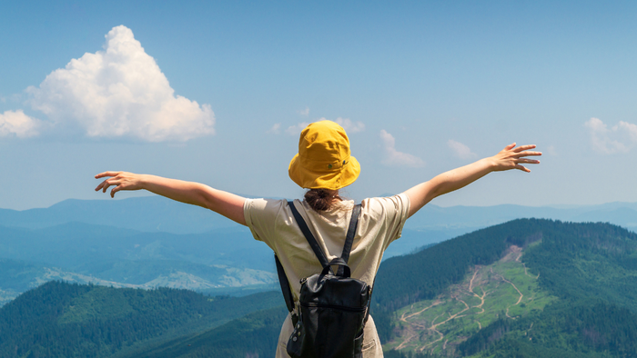 Woman with yellow hat and backpack standing arms open on mountain