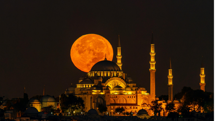 Illuminated mosque at night with a large orange supermoon rising behind it