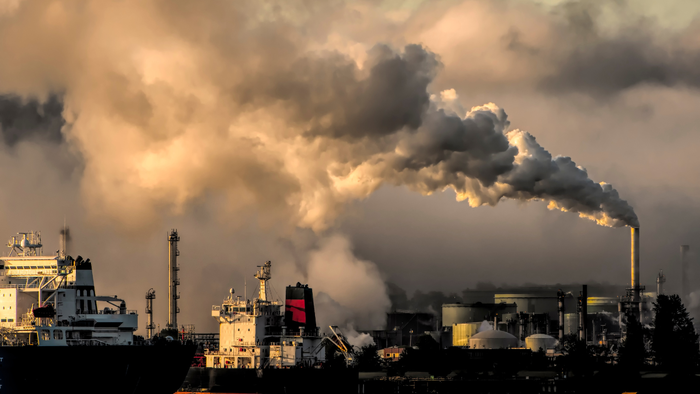 Industrial chimneys and ships emitting thick smoke at sunset, symbolising air pollution near a port.