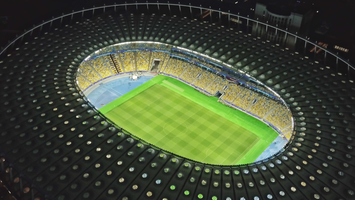 Aerial view of a fully lit football stadium at night, with empty seats and a well-maintained pitch.