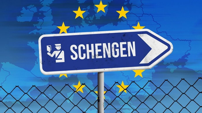 Blue Schengen border sign in front of a map of Europe with yellow EU stars and a wire fence.