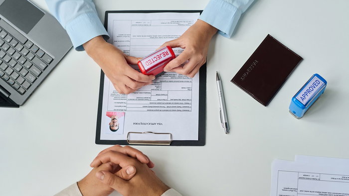 Consulate desk with a Schengen visa application as an officer stamps “REJECTED”; passport and an “APPROVED” stamp visible.