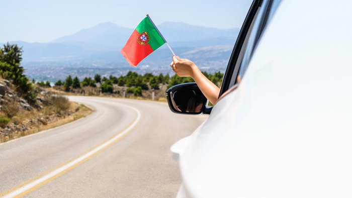 A hand holding a Portuguese flag out of a car window on a rural road in Portugal.