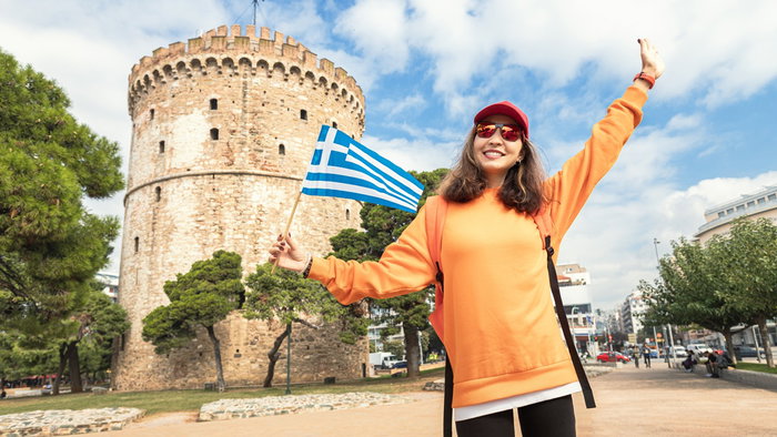 Woman holding a Greek flag in front of the White Tower in Thessaloniki, symbolising residency and relocation to Greece under the Golden Visa programme.