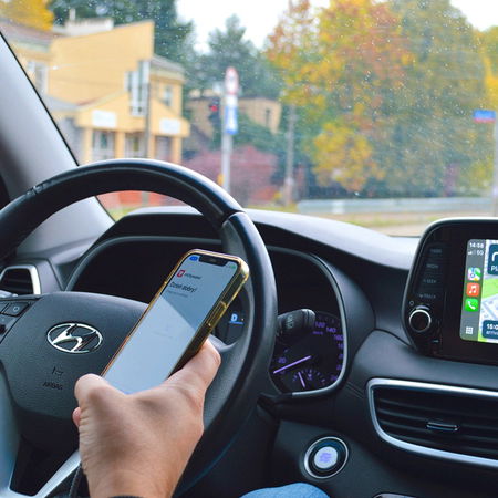 Driver holding a mobile phone while driving with navigation screen visible inside a car.