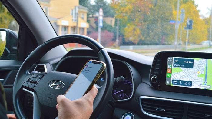 Driver holding a mobile phone while driving with navigation screen visible inside a car.