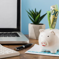 View of a office desk with pen, plant, laptop & piggy bank and a 100€ note in the top