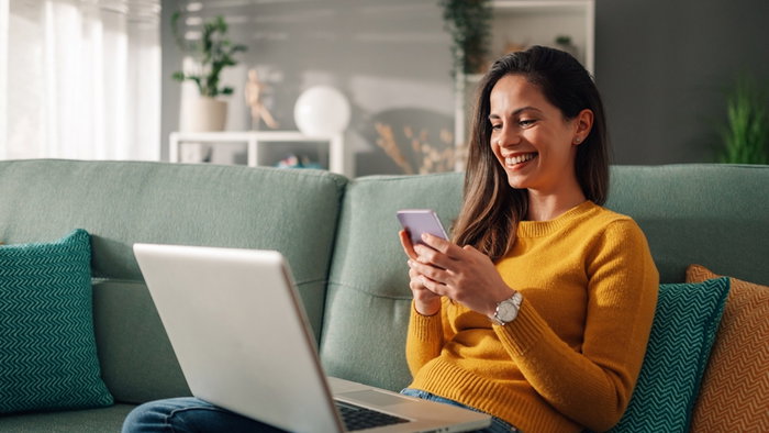 Portrait of a woman using laptop and a phone and checking email or news online while sitting on sofa at home.