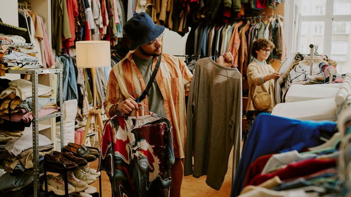 Person browsing racks of clothing in bright thrift store with various garments on display during daylight Peering at different shirts and examining styles and options