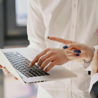 View on a woman holding a laptop in hand whilst typing and another person pointing at the screen