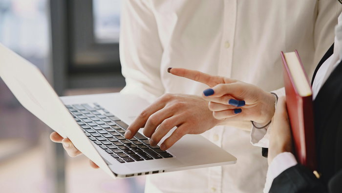 View on a woman holding a laptop in hand whilst typing and another person pointing at the screen