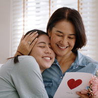 May Mother's day young adult grown up child cuddle hug give flower gift box red heart card to mature middle aged mum. Love kiss care mom asia people sitting at home sofa happy smile enjoy family time.