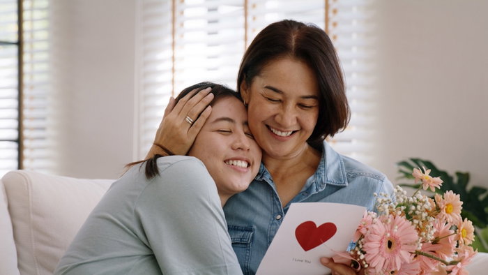 May Mother's day young adult grown up child cuddle hug give flower gift box red heart card to mature middle aged mum. Love kiss care mom asia people sitting at home sofa happy smile enjoy family time.