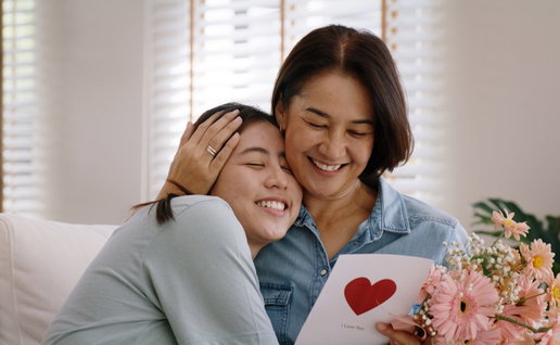 May Mother's day young adult grown up child cuddle hug give flower gift box red heart card to mature middle aged mum. Love kiss care mom asia people sitting at home sofa happy smile enjoy family time.