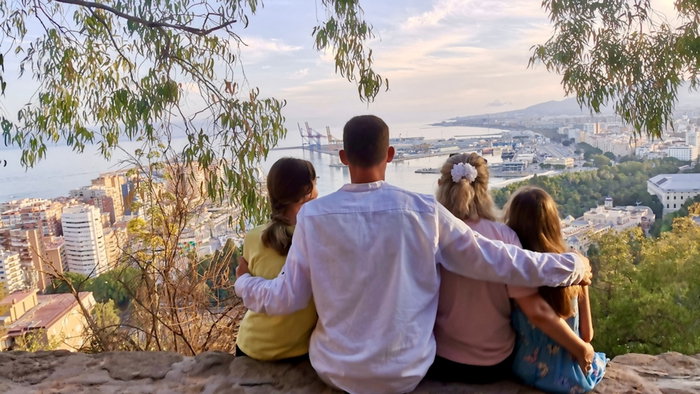Rear view of a family sitting together on a hilltop viewpoint, enjoying the panoramic view of Malaga city and the Mediterranean Sea, Spain, on a sunny day.