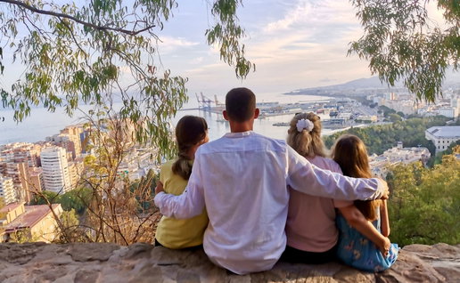 Rear view of a family sitting together on a hilltop viewpoint, enjoying the panoramic view of Malaga city and the Mediterranean Sea, Spain, on a sunny day.