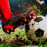 A low-angle action shot capturing the moment a red-socked foot connects with a dirty soccer ball, kicking up dirt and grass during intense game play.