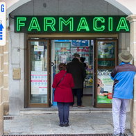 A street-level view of a pharmacy entrance under a stone archway with a bright green "FARMACIA" neon sign. A woman in a red coat and a man in a blue jacket stand outside the glass doors, while another person enters. To the left, a white parking sign indicates "24 Horas."
