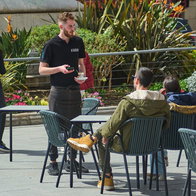 A waiter in a black uniform and apron serves a small glass of coffee to a customer at a sunny outdoor café terrace in a Spanish plaza, representing the local hospitality industry and labor market.