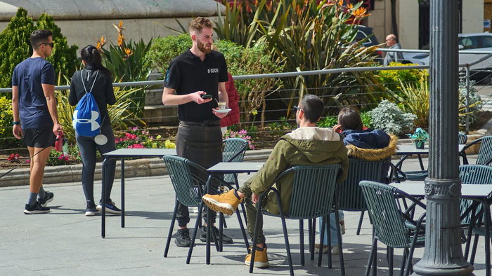 A waiter in a black uniform and apron serves a small glass of coffee to a customer at a sunny outdoor café terrace in a Spanish plaza, representing the local hospitality industry and labor market.
