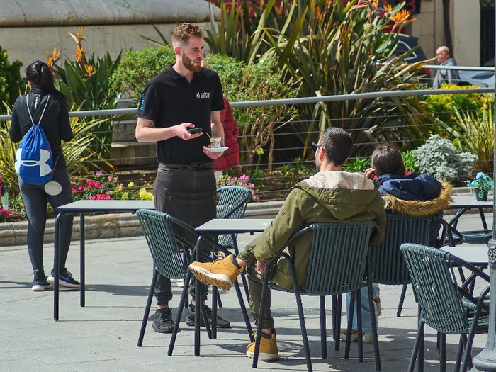 A waiter in a black uniform and apron serves a small glass of coffee to a customer at a sunny outdoor café terrace in a Spanish plaza, representing the local hospitality industry and labor market.