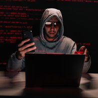 Hooded hacker using a smartphone and laptop in a dark room during a cyberattack linked to phishing and online scams.