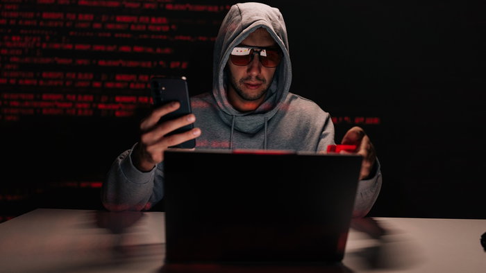 Hooded hacker using a smartphone and laptop in a dark room during a cyberattack linked to phishing and online scams.