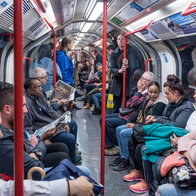 A wide-angle, interior view of a crowded London Underground train carriage with passengers seated and standing along the aisle, featuring bright overhead lighting and iconic red handrails.