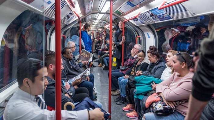 A wide-angle, interior view of a crowded London Underground train carriage with passengers seated and standing along the aisle, featuring bright overhead lighting and iconic red handrails.