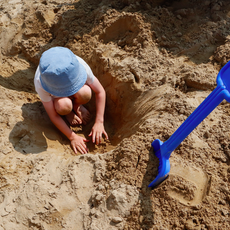 Child digging in the sand on the beach with a blue shovel stuck in the sand