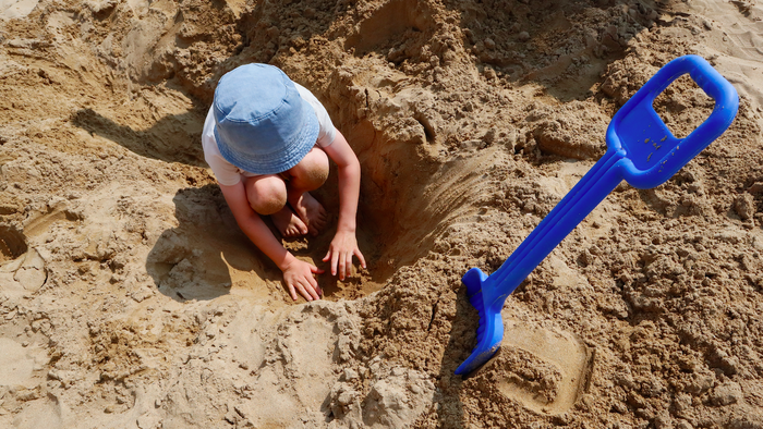 Child digging in the sand on the beach with a blue shovel stuck in the sand