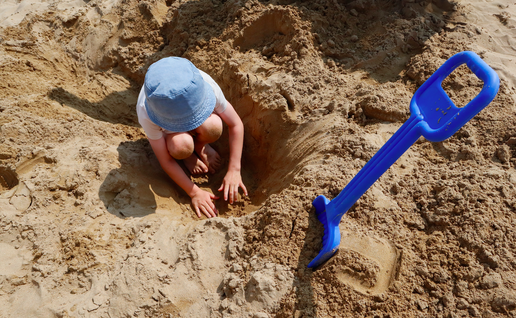 Child digging in the sand on the beach with a blue shovel stuck in the sand