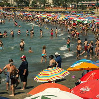 A crowded beach on the Costa del Sol packed with tourists swimming in the sea and sunbathing under colorful umbrellas on a bright, sunny day.