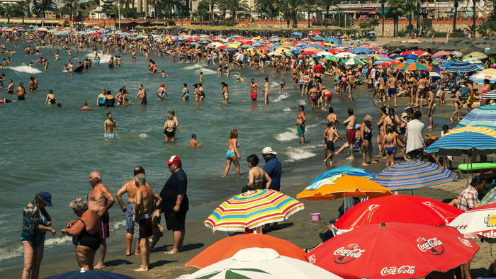 A crowded beach on the Costa del Sol packed with tourists swimming in the sea and sunbathing under colorful umbrellas on a bright, sunny day.