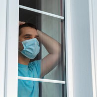 Home quarantine, self-isolation because of the Coronavirus disease, COVID-19. Man in a medical mask near the window. Boredom and depression during quarantine. Waiting for quarantine to be canceled.
