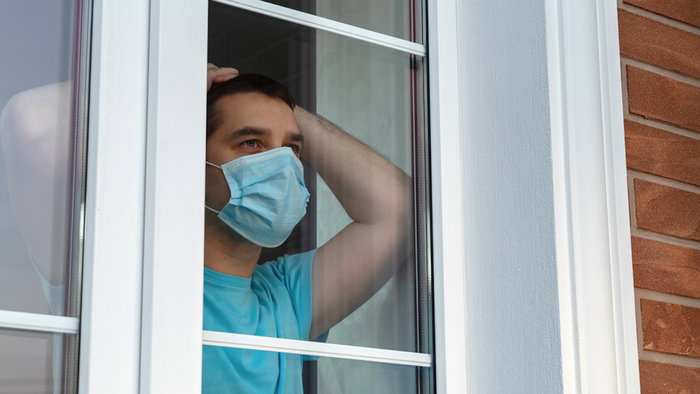 Home quarantine, self-isolation because of the Coronavirus disease, COVID-19. Man in a medical mask near the window. Boredom and depression during quarantine. Waiting for quarantine to be canceled.