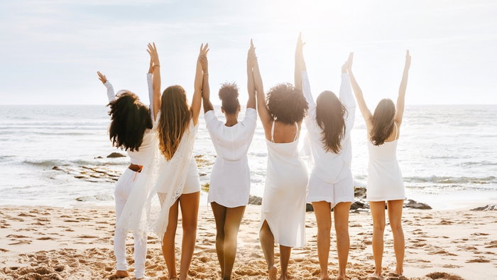 Women on the beach. Back view of group of diverse ladies walking by the seashore, holding and raising hands, enjoying sunset and beautiful ocean view
