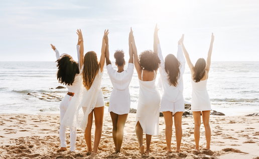 Women on the beach. Back view of group of diverse ladies walking by the seashore, holding and raising hands, enjoying sunset and beautiful ocean view