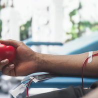 Close Up Shot Of Hand Of Male Blood Donor With an Attached Catheter. Black Man Holding Heart-Shaped Red Ball To Pump Blood Through The Tubing Into Bag. Donation For Organ Transplant Patients.