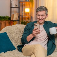 Young man at home starts morning sitting on sofa with tea, reading smartphone with calm focus. Caucasian guy looks peaceful, focused on news and relaxed, enjoying warm drink in quiet home moment.
