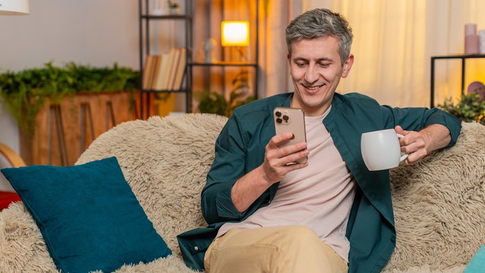 Young man at home starts morning sitting on sofa with tea, reading smartphone with calm focus. Caucasian guy looks peaceful, focused on news and relaxed, enjoying warm drink in quiet home moment.