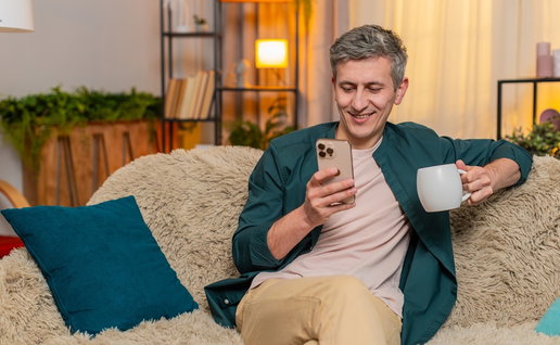 Young man at home starts morning sitting on sofa with tea, reading smartphone with calm focus. Caucasian guy looks peaceful, focused on news and relaxed, enjoying warm drink in quiet home moment.