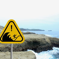 Tsunami warning sign standing beside a rocky Mediterranean coastline with waves hitting the shore.