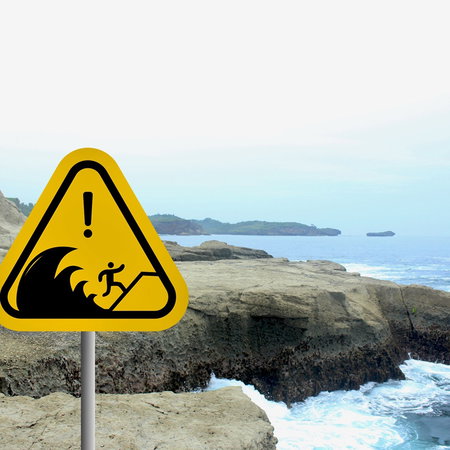 Tsunami warning sign standing beside a rocky Mediterranean coastline with waves hitting the shore.