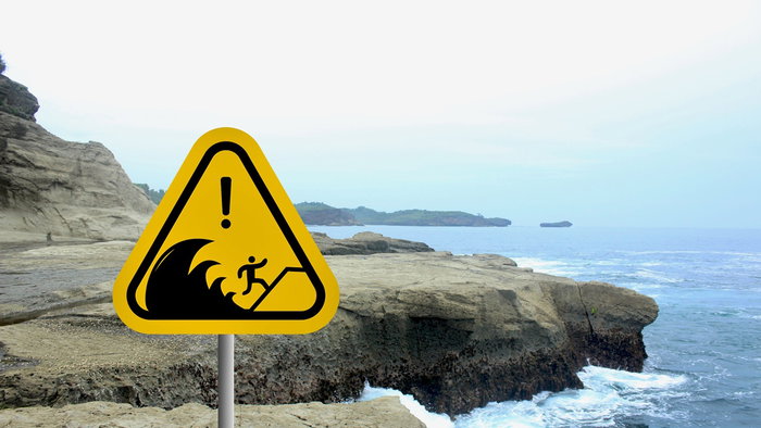 Tsunami warning sign standing beside a rocky Mediterranean coastline with waves hitting the shore.